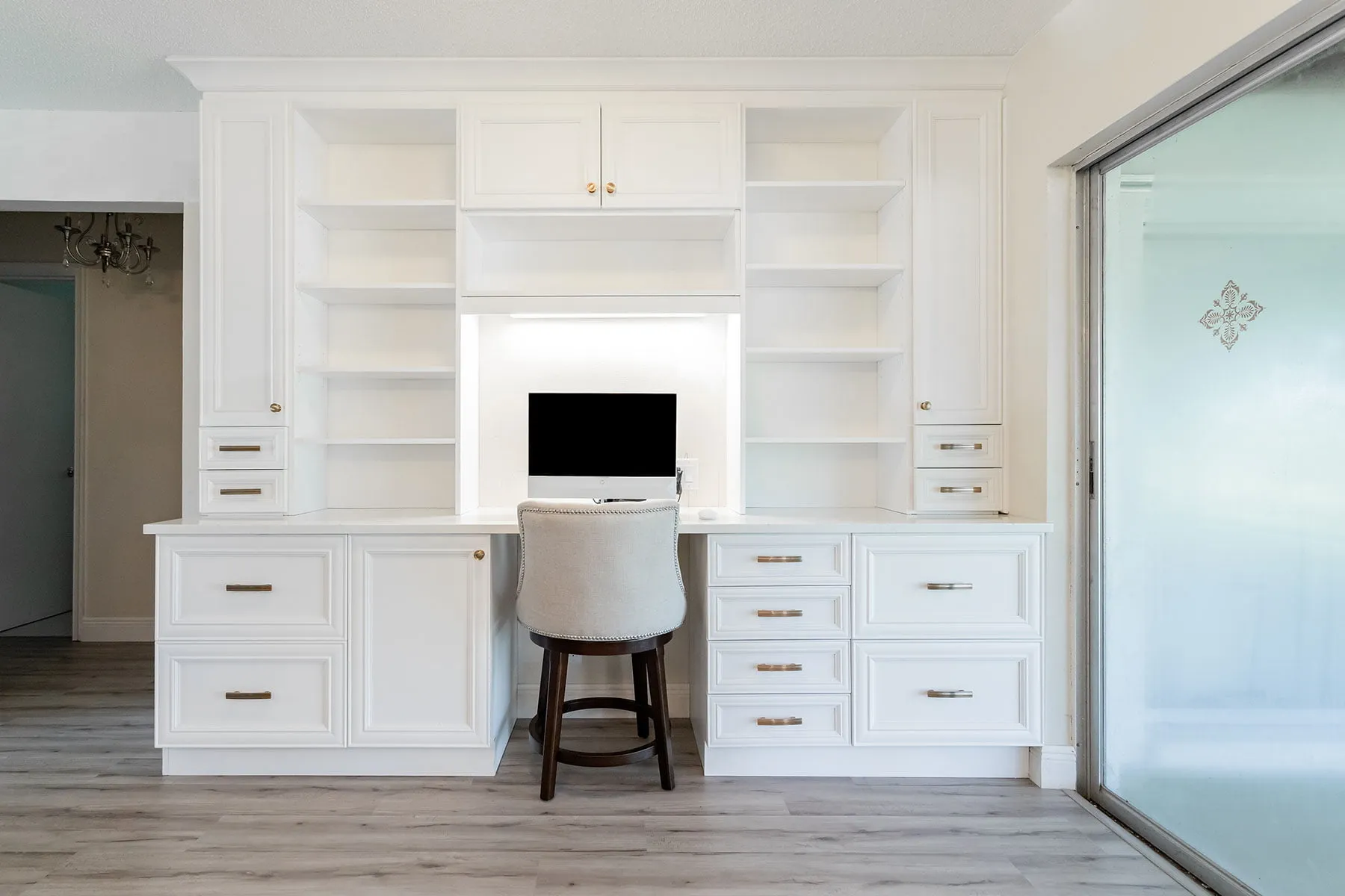 White shaker built-in desk nook with lit open shelving and drawers beside a glass sliding door